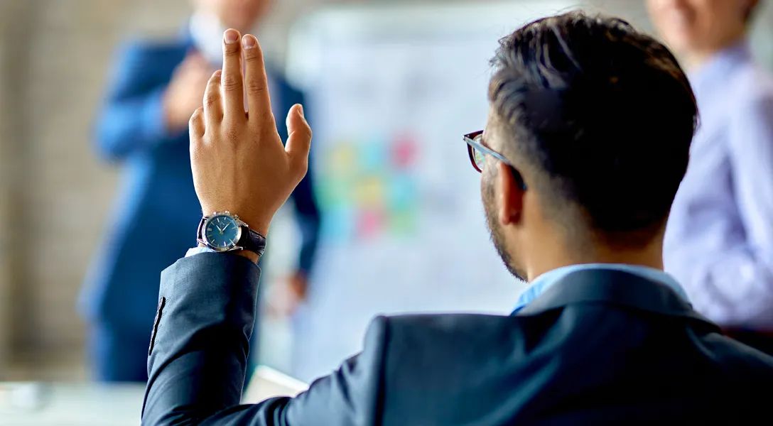 Stock photo of a man with short cropped hair in a business suit raising his hand left hand. He faces towards presenters and also wears a black wrist watch.