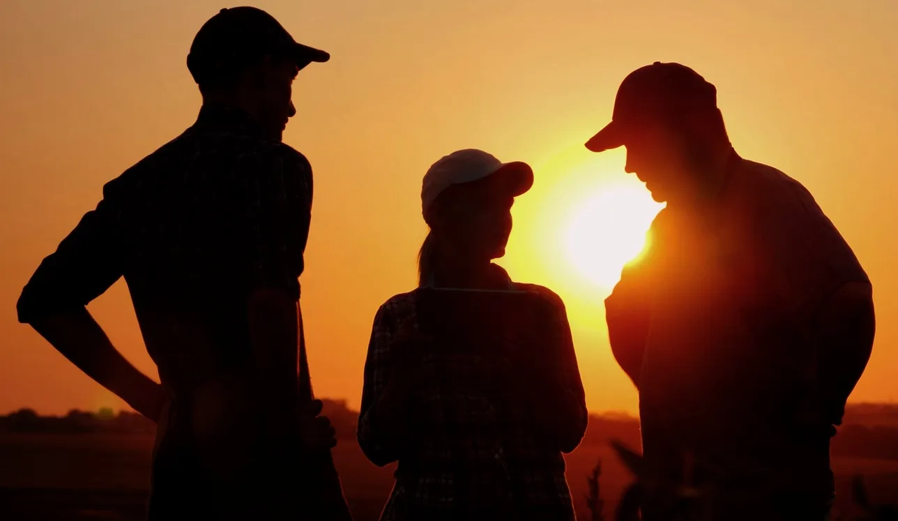 Shadowy silhouette of three individuals wearing hats in a field at sunset.